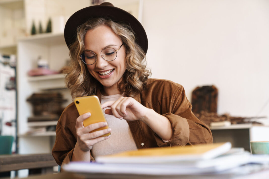 smiling woman eyeglasses hat using cellphone while studying with exercise books home